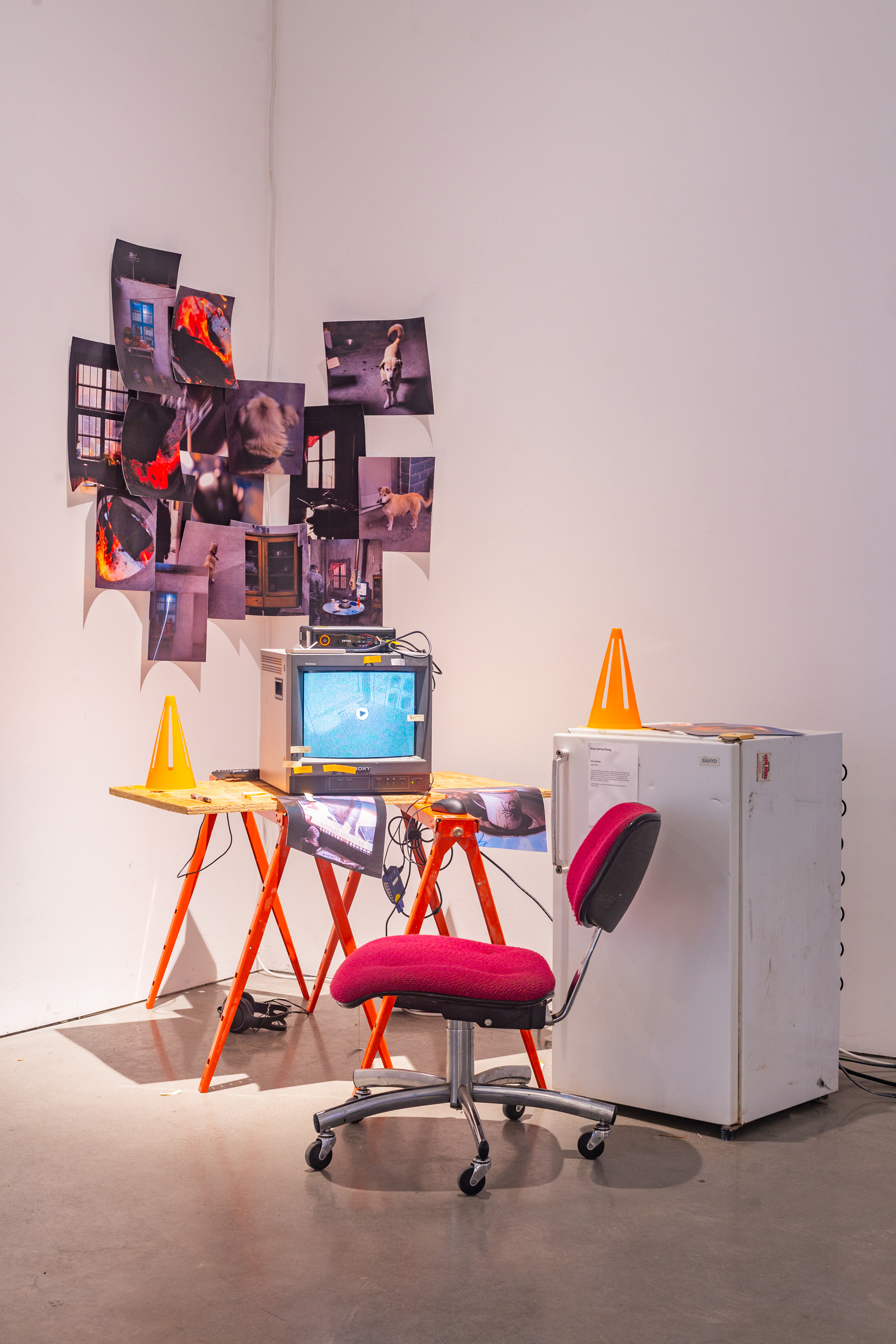 A sculptural piece, on the right is a white fridge with a plastic cone on top of it, in the center is a makeshift construction desk with plywood and an office chair, on top of it is a CRT tv with visuals on it, in the background is printed posters of photogrpahy.